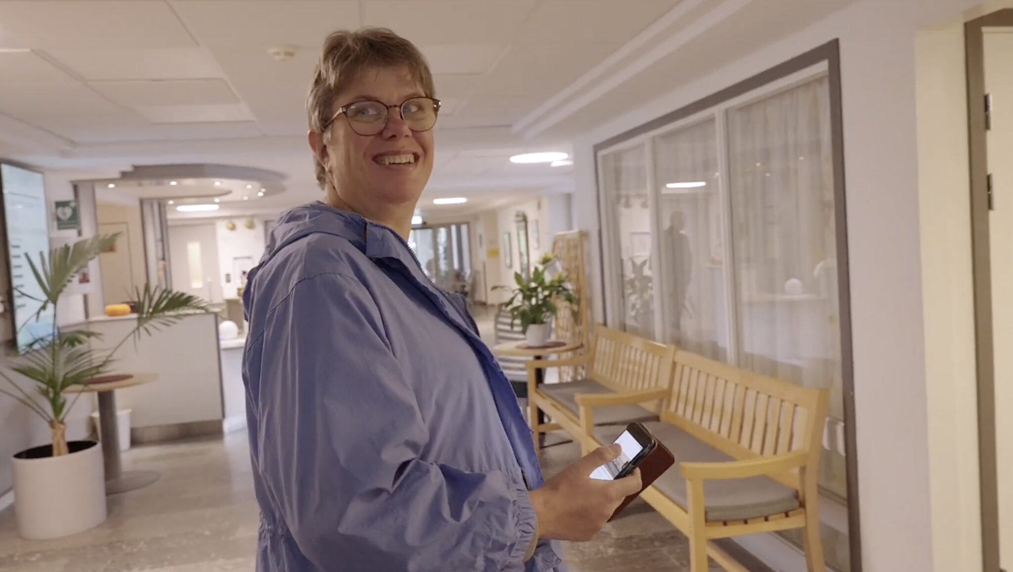 En leende person med glasögon och ljusblå jacka håller i en telefon medan han går längs en ljus korridor med träbänkar och växter.