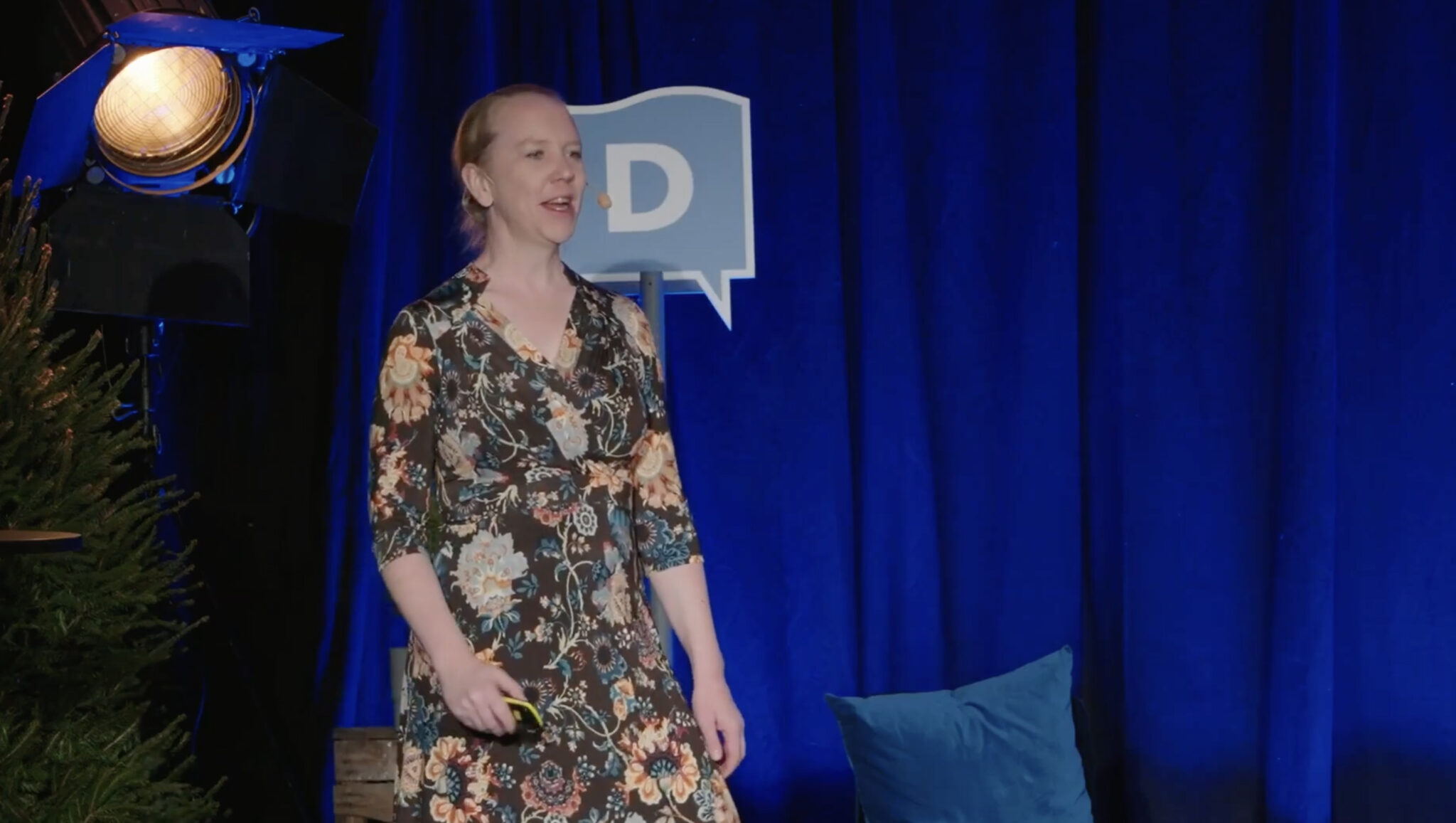 En person i blommig klänning står på scenen framför blå gardiner och talar. I bakgrunden finns en stor scenlampa, en blå kudde och en skylt med bokstaven D.