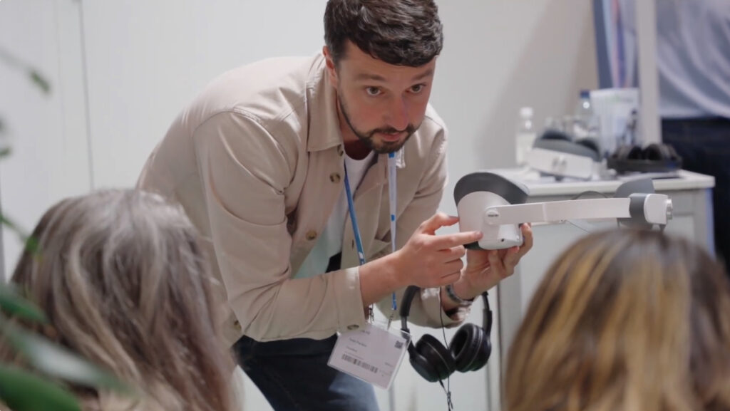 En man med en konferensbricka demonstrerar ett VR-headset för två sittande personer på ett evenemang inomhus. Han pekar på enheten medan de andra tittar uppmärksamt på.