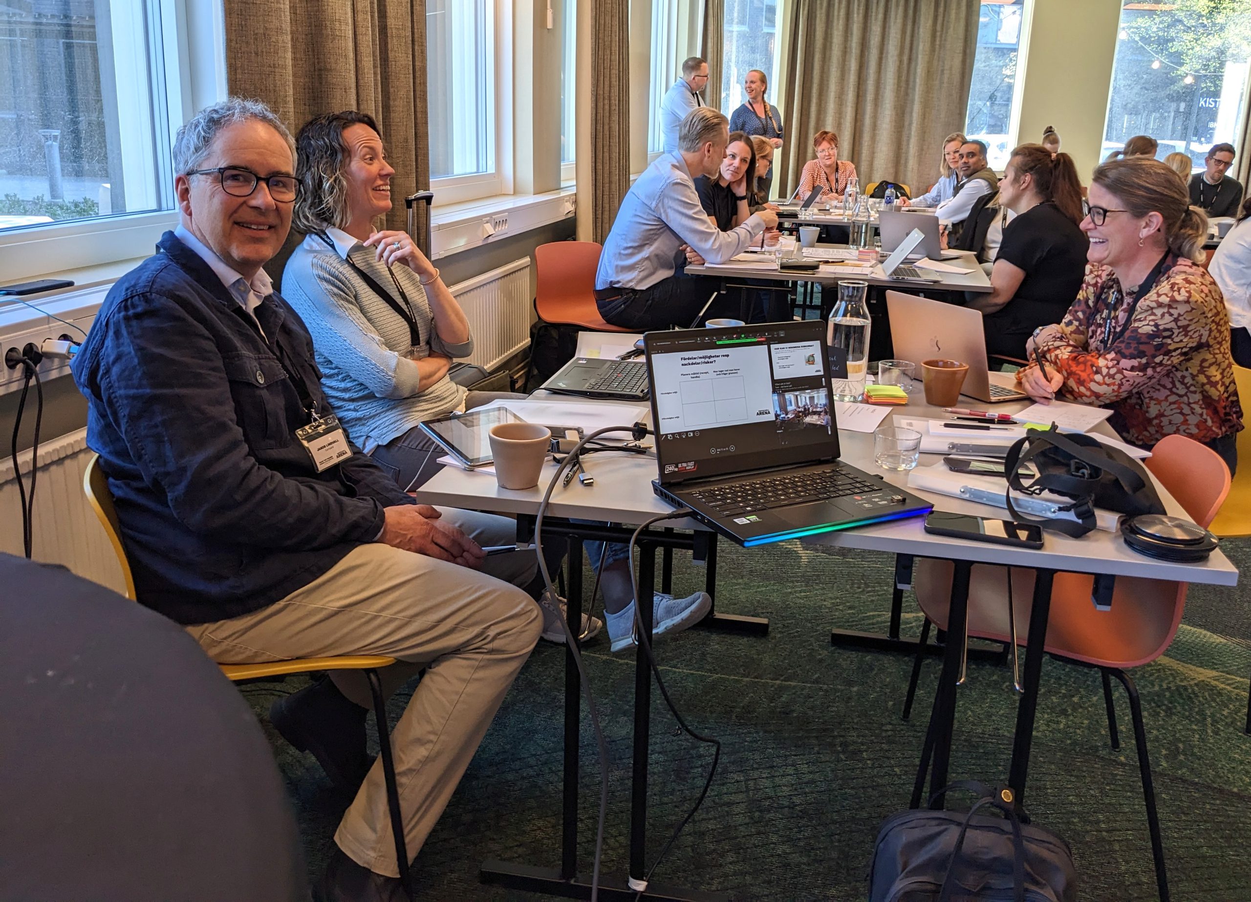 En grupp människor sitter vid bord i ett ljust rum, engagerade i samtal och arbetar på bärbara datorer under en workshop eller ett möte. Tre personer i förgrunden ler, medan andra arbetar i bakgrunden.