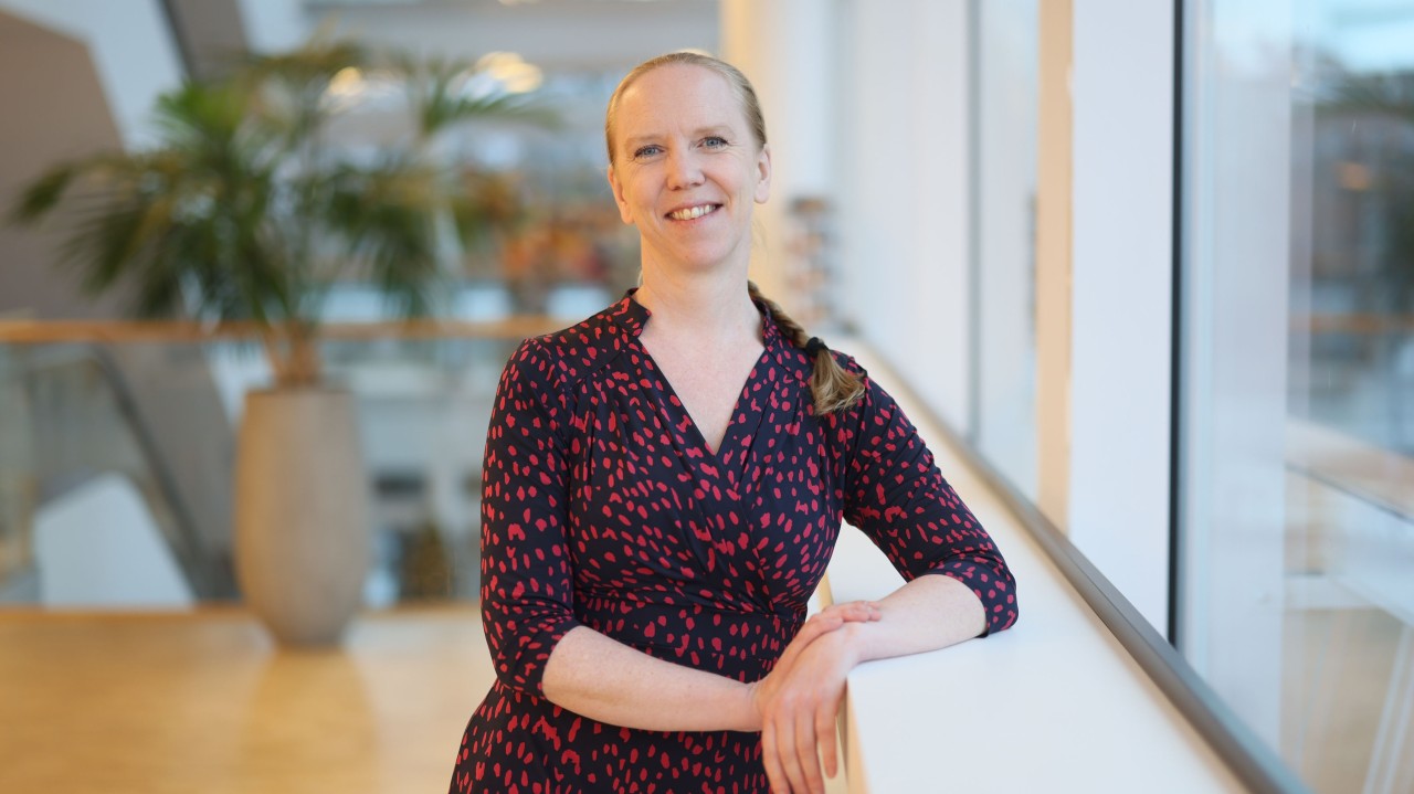 En kvinna med blont hår i en fläta, klädd i en mörk klänning med rött mönster, står leende och lutar sig mot ett räcke inomhus intill stora fönster med en krukväxt i bakgrunden.