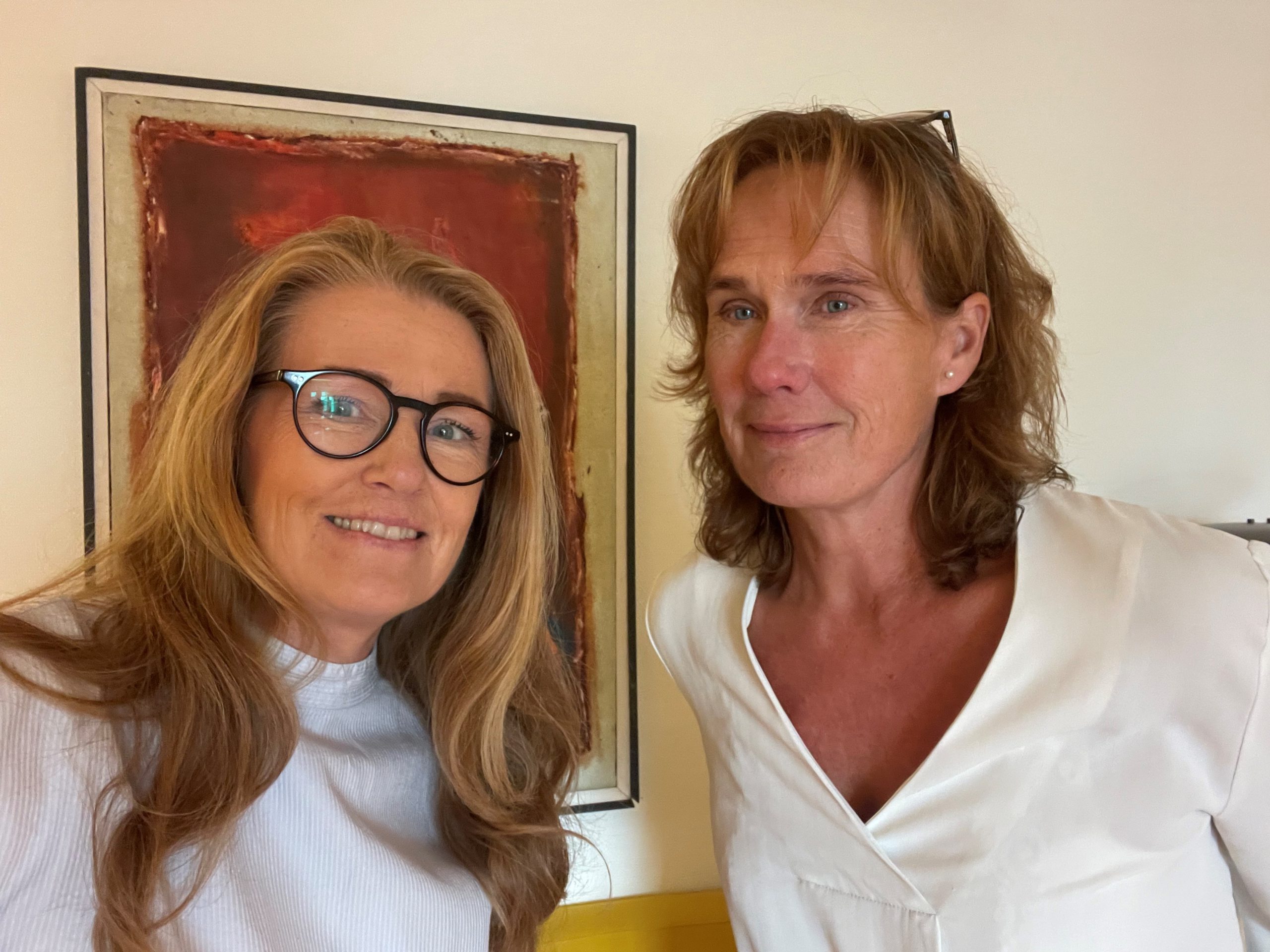 Två kvinnor med långt hår står inomhus och ler mot kameran. Den ena bär glasögon och en ljus topp, medan den andra bär en vit skjorta. En inramad abstrakt målning med röda toner hänger på väggen bakom dem.