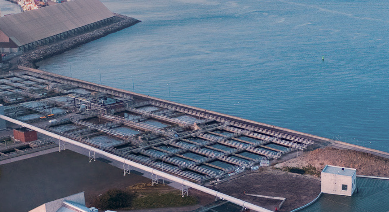 Flygfoto över en stor vattenreningsanläggning vid kusten, med flera rektangulära sedimenteringsbassänger och industribyggnader intill ett vattendrag. En betongbarriär skiljer anläggningen från havet.