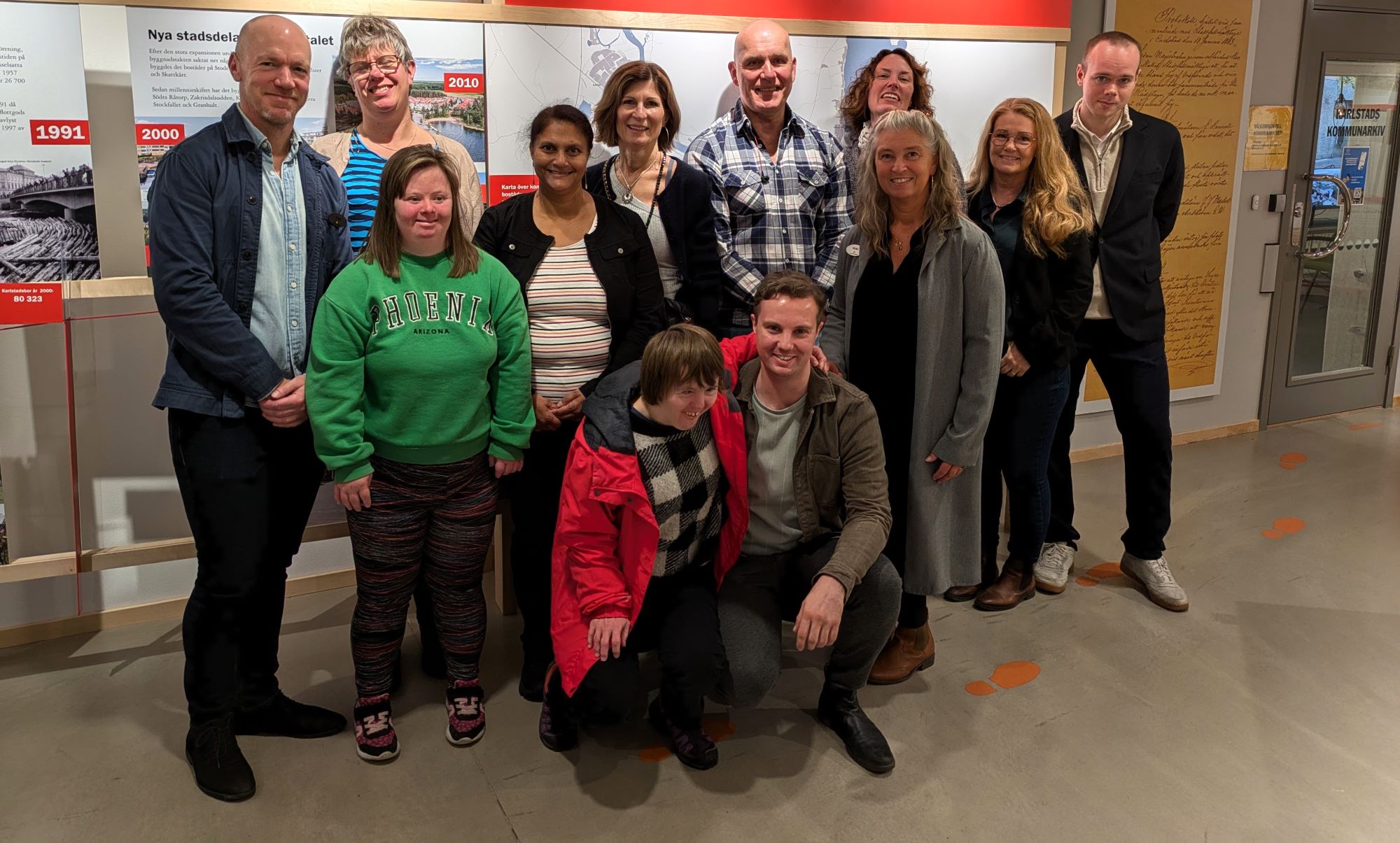 En grupp på tretton personer poserar och ler tillsammans inomhus framför en utställning med historiska informationstavlor på väggen. De flesta står upp och några knäböjer längst fram.