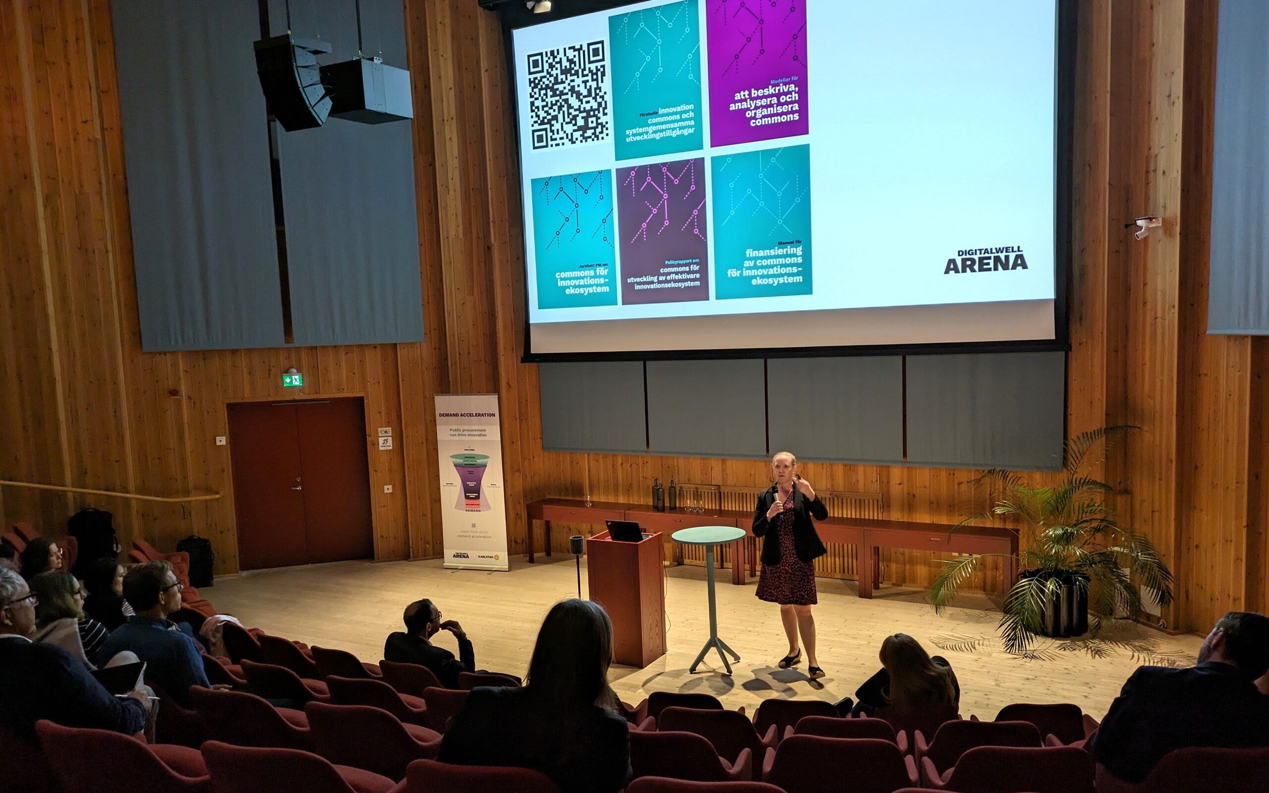 En kvinna står på scenen och håller en presentation i en föreläsningssal i trä. Hon talar i en mikrofon och bakom henne projiceras bilder med QR-koder och färgglad grafik. Åhörarna sitter i röda stolar vända mot henne.