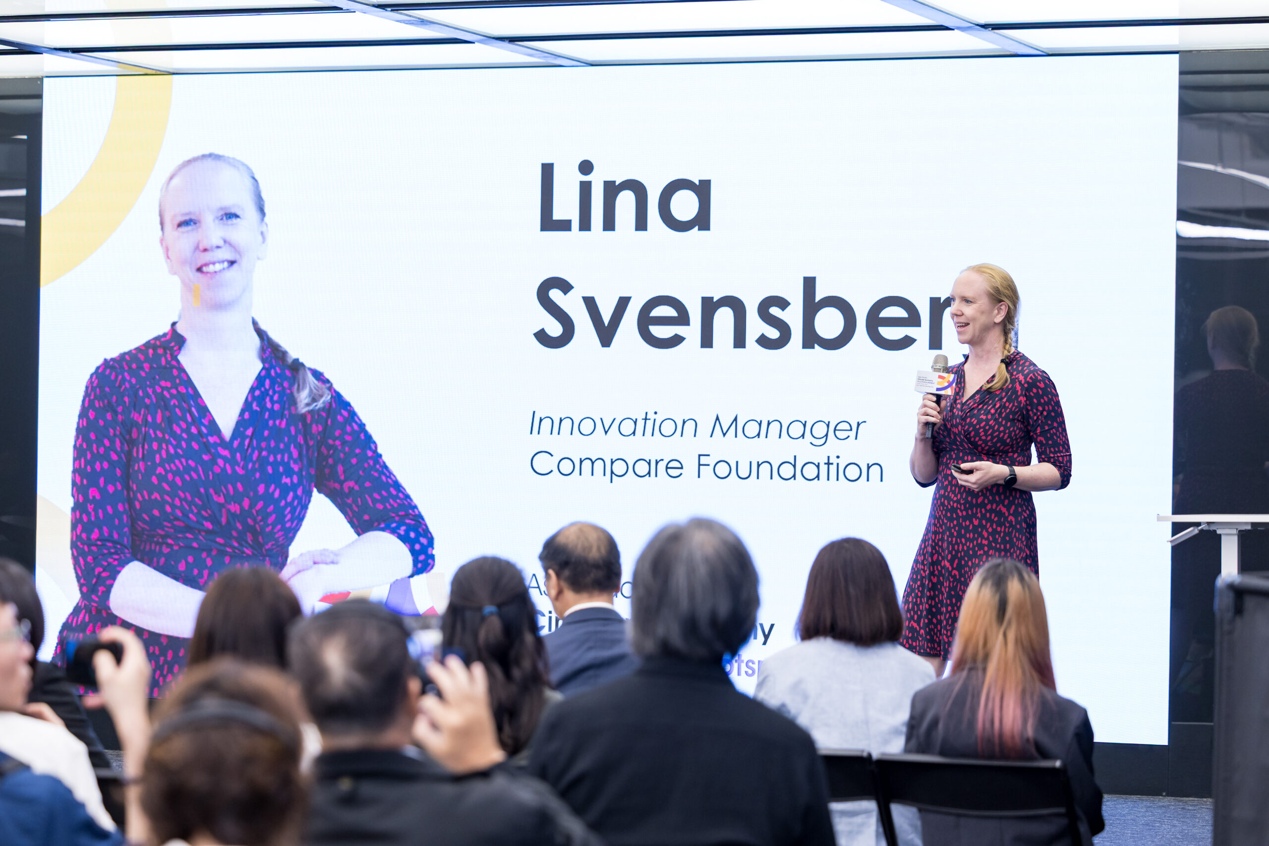 En kvinna i mönstrad klänning talar på scen med mikrofon inför publik. Bakom henne finns en stor skärm som visar hennes foto och text: Lina Svensber, innovationschef, Stiftelsen Compare.