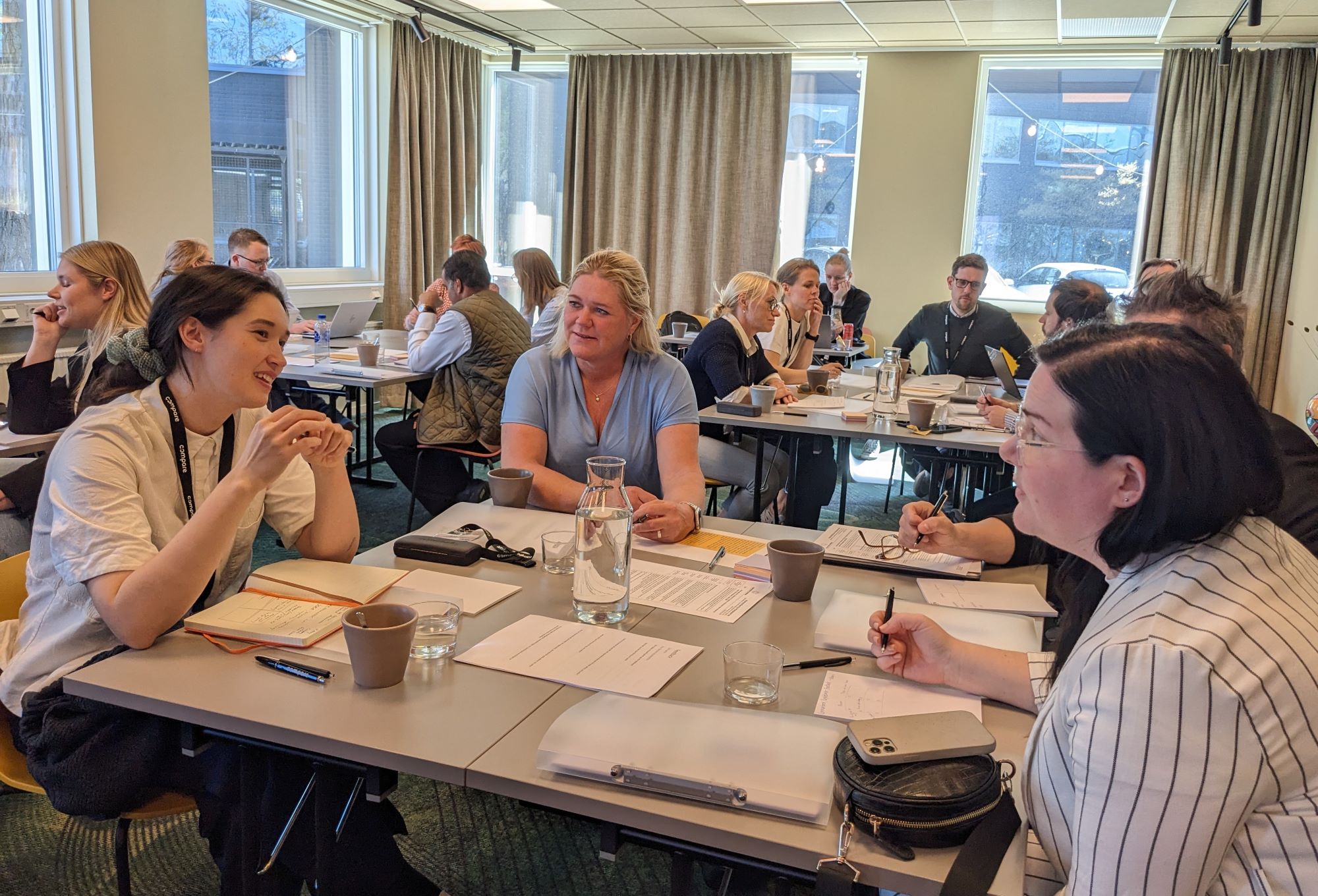Människor sitter i grupper runt bord i ett ljust konferensrum och pratar och antecknar. Papper, anteckningsblock och koppar står på borden och stora fönster släpper in naturligt ljus.