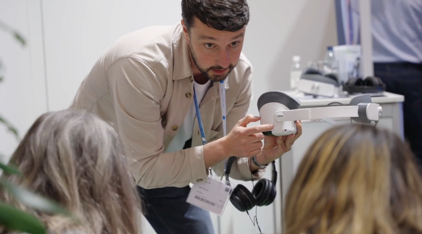 En man demonstrerar en handhållen enhet för en liten grupp och pekar på dess funktioner. Han bär namnskylt och jacka och två personer med långt hår sitter i förgrunden och tittar uppmärksamt på.