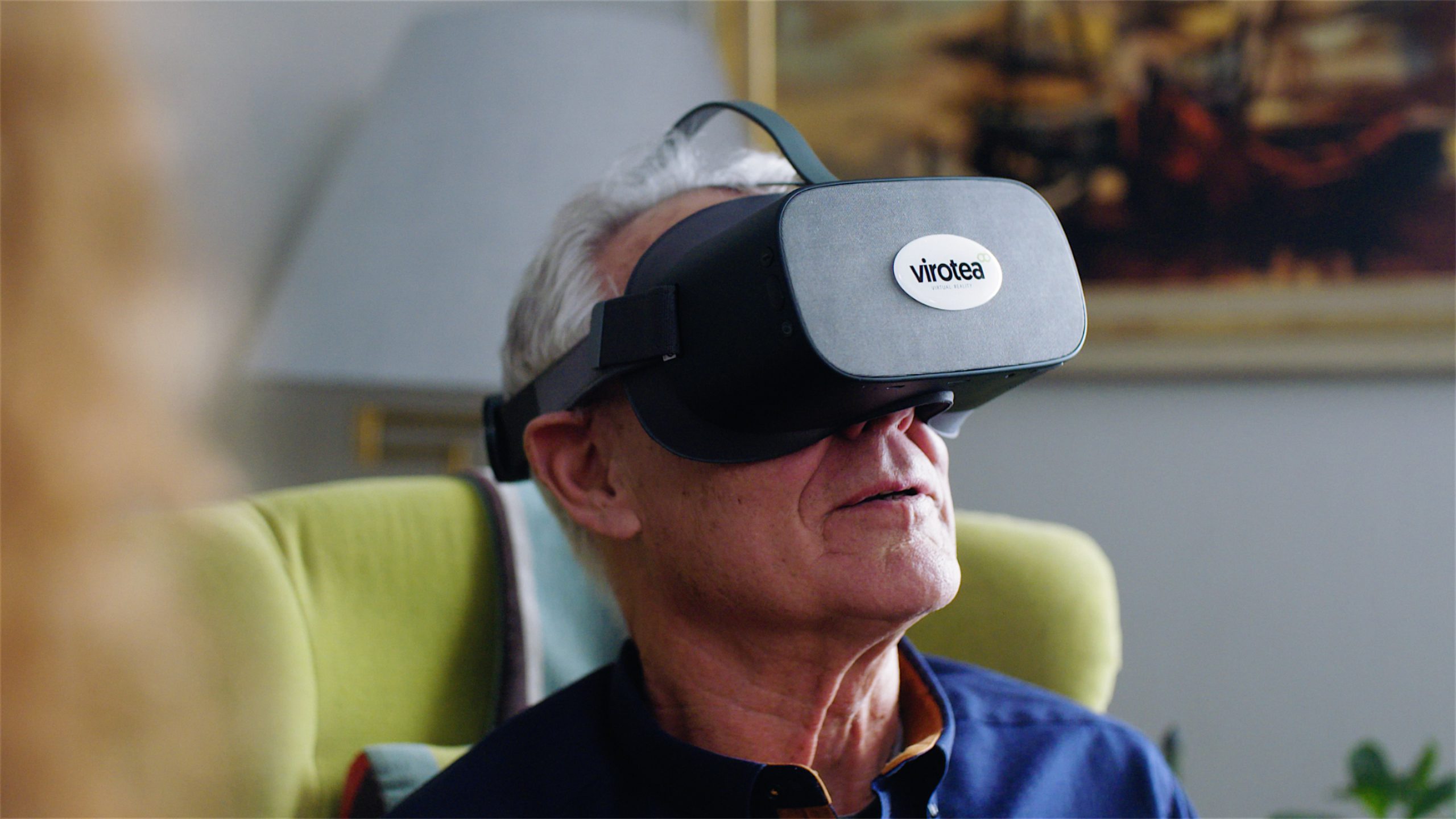 En äldre man sitter i en grön stol med ett virtual reality-headset av märket Virotea och är uppslukad av en VR-upplevelse. Bakgrunden är mjukt upplyst med en målning på väggen.