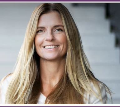 En kvinna med långt, rakt blont hår ler mot kameran. Hon har på sig en ljus topp och står framför en suddig bakgrund av vita trappsteg.