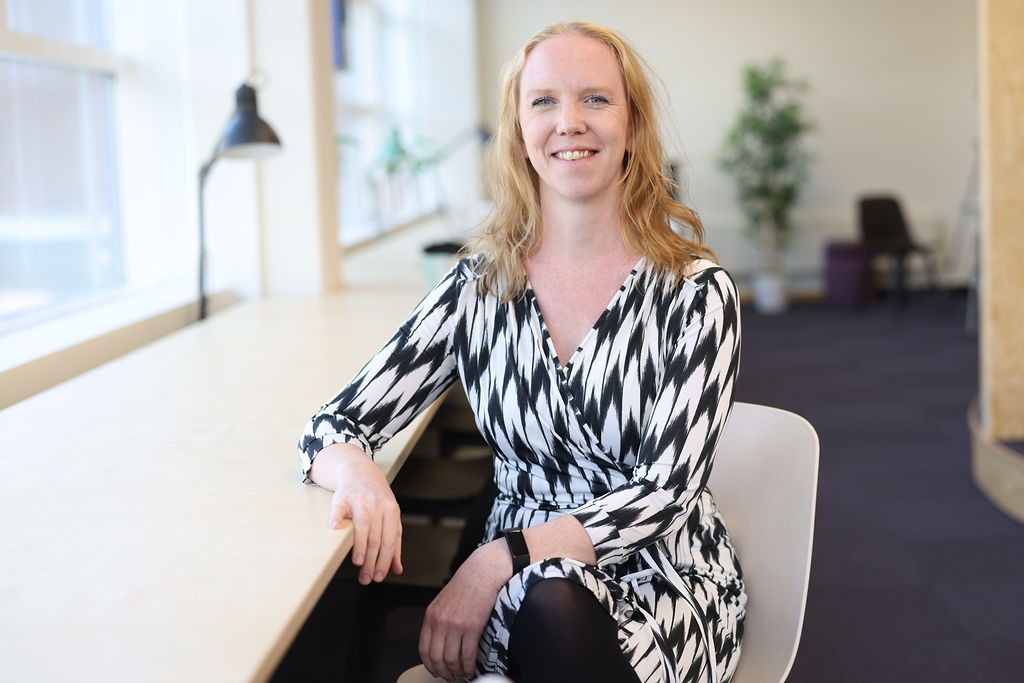 En kvinna med långt blont hår sitter vid ett skrivbord i ett ljust kontor och ler mot kameran. Hon bär en svartvit mönstrad klänning. I bakgrunden syns stora fönster, en lampa och växter.