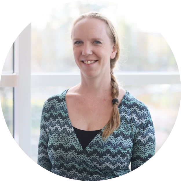 En kvinna med ljus hy och blont hår i en sidfläta ler mot kameran. Hon har på sig en mönstrad grön och svart topp och står framför ett ljust fönster.